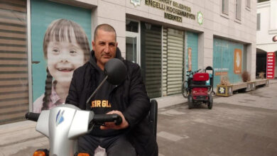 Görselde, bir adam elektrikli engelli aracı üzerinde oturuyor ve kameraya bakıyor. Arkasında "Aksaray Belediyesi Engelli Koordinasyon Merkezi" tabelası bulunan bir bina yer alıyor. Binanın camlarında engelli bireylerle ilgili görseller ve yazılar bulunmakta. Arka planda bir başka engelli aracı park halinde duruyor.