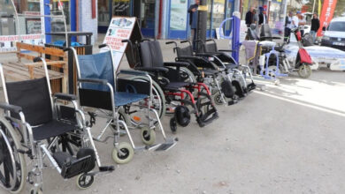 Görselde bir dizi tekerlekli sandalye, hasta taşıma lifti, akülü araçlar ve hasta yatakları açık havada sergileniyor. Ekipmanlar kaldırım kenarında sıralanmış ve çeşitli türlerde manuel ve akülü tekerlekli sandalyeler yer alıyor. Arkada bazı dükkanlar ve birkaç kişi görülüyor.