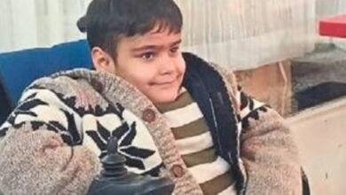 Fotoğrafta, genç bir çocuk akülü tekerlekli sandalyesinde oturuyor. Üzerinde beyaz, siyah desenli kalın bir hırka ve altına yeşil-beyaz çizgili bir kazak giymiş. Hafifçe gülümseyerek kameraya bakıyor. Arka planda perde ve pencere bulunan bir kapalı mekân yer alıyor.