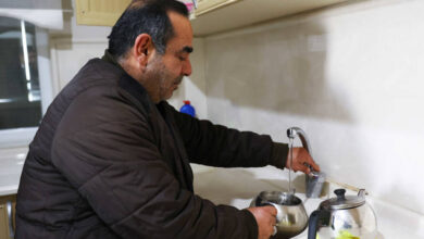 Görselde, orta yaşlı bir adam mutfakta su dolu bir çaydanlığı musluktan dolduruyor. Üzerinde kahverengi kalın bir mont bulunuyor. Tezgâhta bir demlik ve bir bez yer alıyor. Mutfak temiz ve düzenli görünüyor, üst dolaplar açık renk ahşap kaplamaya sahip.