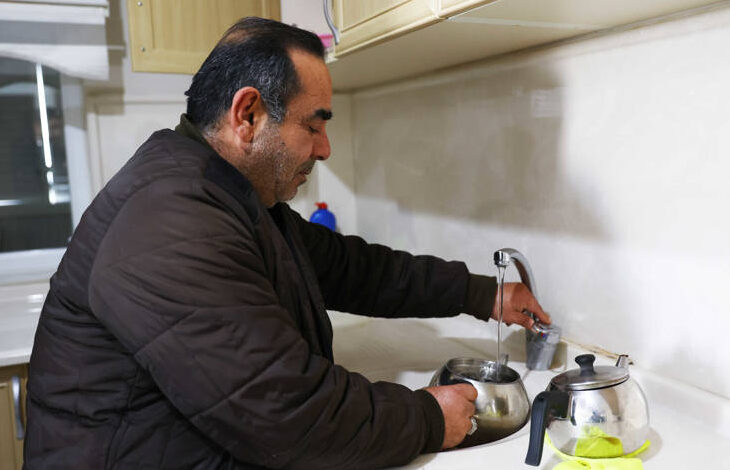 Görselde, orta yaşlı bir adam mutfakta su dolu bir çaydanlığı musluktan dolduruyor. Üzerinde kahverengi kalın bir mont bulunuyor. Tezgâhta bir demlik ve bir bez yer alıyor. Mutfak temiz ve düzenli görünüyor, üst dolaplar açık renk ahşap kaplamaya sahip.