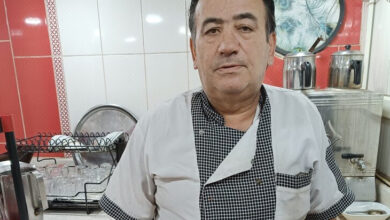 Bu görselde, aşçı üniforması giymiş bir adam mutfakta poz veriyor. Arkada çay bardakları, çaydanlıklar ve mutfak eşyaları görülebiliyor.