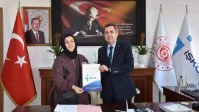 Fotoğrafta, bir kadın ve bir erkek resmi bir ofiste bulunuyor. Erkek takım elbise giymiş ve kadına üzerinde "İŞKUR" logosu bulunan bir dosya veriyor. Kadın başörtülü, elinde çanta tutuyor ve dosyayı alırken gülümsüyor. Arkada Türkiye bayrağı, İŞKUR ve Çalışma ve Sosyal Güvenlik Bakanlığı flamaları, Cumhurbaşkanı Recep Tayyip Erdoğan'ın bir fotoğrafı ve Atatürk'ün bir portresi yer alıyor.