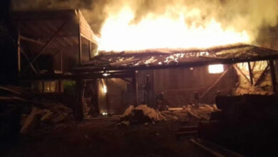 Görselde, gece vakti alevler içinde kalan bir ahşap yapı görülüyor. Yangın şiddetli bir şekilde devam ederken, yoğun duman gökyüzüne yükseliyor. Binanın büyük kısmı çökmüş veya zarar görmüş durumda, etrafta dağılmış ahşap malzemeler bulunuyor. Ortam karanlık olup yangının parlak alevleri çevreyi aydınlatıyor.