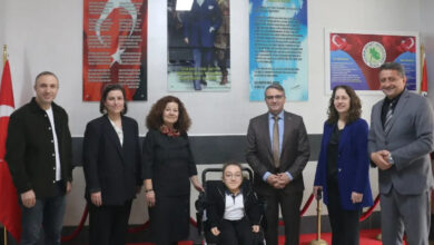 Görselde, bir bina içinde düzenlenen resmi bir etkinlikte bir grup insan poz veriyor. Ortada akülü tekerlekli sandalyede oturan genç bir birey bulunuyor. Etrafında ise resmi kıyafetler giymiş altı yetişkin yer almakta. Arkada Atatürk'ün fotoğrafı ve çeşitli yazılar içeren panolar, ayrıca Türk bayrakları bulunuyor.