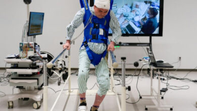 Bu fotoğraf, Çin'in geliştirdiği nöral baypas teknolojisini gösteriyor. Görselde, beyaz bir önlük içinde, başında elektrotlar bulunan bir hasta, mavi destek kemeriyle yürümeye çalışıyor. Yanında tıbbi cihazlar, bilgisayar sistemleri ve hasta takibini gösteren bir ekran yer alıyor.
