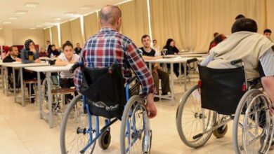 Görselde, bir sınıf ortamında bulunan öğrenciler görülüyor. Ön planda iki tekerlekli sandalye kullanan birey, sırtı kameraya dönük şekilde oturuyor. Arkada, masa ve sandalyelerde oturan diğer öğrenciler bulunuyor. Sınıfın aydınlık ve geniş bir alan olduğu, perdeyle kapatılmış büyük pencerelerin bulunduğu dikkat çekiyor.