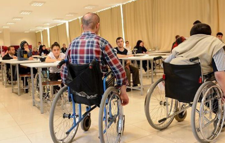 Görselde, bir sınıf ortamında bulunan öğrenciler görülüyor. Ön planda iki tekerlekli sandalye kullanan birey, sırtı kameraya dönük şekilde oturuyor. Arkada, masa ve sandalyelerde oturan diğer öğrenciler bulunuyor. Sınıfın aydınlık ve geniş bir alan olduğu, perdeyle kapatılmış büyük pencerelerin bulunduğu dikkat çekiyor.