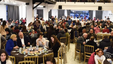 Görselde büyük bir salonda düzenlenmiş bir iftar programı görülüyor. Altın rengi sandalyeler ve kahverengi örtülü yuvarlak masalar etrafında çok sayıda insan oturuyor ve yemek yiyor. Çoğunluğu aileler ve çeşitli yaş gruplarından bireyler oluşturuyor. Arkada perdeyle kapatılmış büyük pencereler ve bir balkon katı var, burada da oturan ve ayakta duran kişiler bulunuyor.