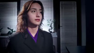 Görselde, genç bir kadın resmi bir ortamda oturuyor. Siyah bir ceket ve içinde mor bir bluz giyiyor. Arkasında panjurları kapalı pencereler ve masanın üzerinde küçük süs bitkileri bulunuyor. Ortam loş ve ciddi bir hava taşıyor. Görüntü, epilepsiye yönelik bir kamu spotundan alınmış olabilir ve bilinçlendirme amacı taşıyan bir sahneye ait gibi görünüyor.