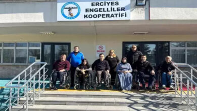 Görselde, Erciyes Engelliler Kooperatifi binasının önünde bir grup insan poz veriyor. Ön sırada tekerlekli sandalyede oturan bireyler, arkada ise ayakta duran kişiler bulunuyor. Binanın girişinde engelsiz erişim için rampalar mevcut. Üst tarafta kooperatifin adının yazılı olduğu bir tabela asılı.