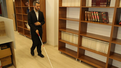 Görselde, elinde beyaz baston taşıyan görme engelli bir adam, bir kütüphane veya çalışma alanı gibi görünen bir ortamda yürümektedir. Siyah bir takım elbise giymektedir ve çevrede ahşap kitap rafları bulunmaktadır. Rafların bazıları kitaplarla doluyken bazıları boştur. Ortam düzenli ve aydınlıktır.