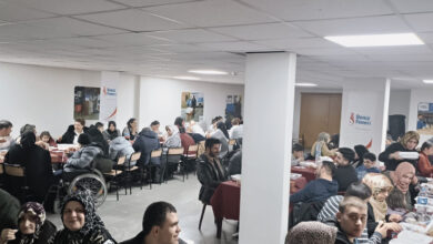 Fotoğrafta, büyük bir salonda düzenlenen toplu bir iftar yemeği görülüyor. Masalar etrafında çok sayıda insan oturmuş, yemeklerini yiyor veya sohbet ediyor. Katılımcılar arasında engelli bireyler de bulunuyor; bir kişi tekerlekli sandalyede oturuyor. Duvarlarda "Deniz Feneri" derneğinin afişleri yer alıyor.