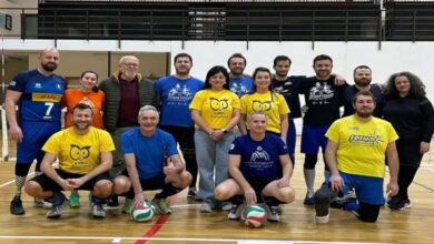 Görselde, bir spor salonunda toplanmış bir grup insan yer alıyor. Grup, voleybol sahasında poz veriyor ve çoğu spor kıyafetleri giymiş. Bazıları sarı, mavi ve siyah tişörtler giymiş olup tişörtlerde çeşitli logolar ve yazılar bulunuyor. Önde çömelmiş halde duran bazı kişiler ellerinde voleybol topları tutuyor. Arkada ise ayakta duran diğer takım üyeleri ve antrenör olabilecek kişiler var. Mekân kapalı bir spor salonu olup, voleybol filesi ve sahadaki çizgiler belirgin şekilde görünüyor.