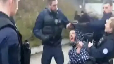 Görselde, birkaç polis memuru bir kişiyi sert bir şekilde gözaltına alıyor. Tekerlekli sandalyede oturan bir adam, bir polis memuru tarafından yumruklanıyor gibi görünüyor. Adamın yüzündeki ifade acı ve şaşkınlık içeriyor. Çevrede başka polis memurları da var ve olay sokakta, açık alanda gerçekleşiyor.