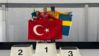Görselde, bir atletizm şampiyonasında ödül töreni anı yer alıyor. Üç sporcu, madalyalarıyla birlikte kürsüde duruyor. Birinci sırada yer alan Türk sporcu kırmızı milli takım forması giymiş ve elinde Türk bayrağını tutuyor. Solunda ikinci olan sporcu ve sağında üçüncü olan sporcu kendi ülkelerinin bayraklarını taşıyor. Arka planda kapalı bir spor salonu ortamı ve fileler bulunuyor. Sporcuların yüzlerinde mutluluk ve gurur ifadesi var.