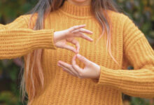 Görselde, hardal sarısı kalın örgü kazak giymiş bir kişi yer alıyor. Kişinin yüzü görünmüyor, ancak uzun, açık kahverengi ve uçları açık renk tonlarına sahip saçları dikkat çekiyor. Ellerini kullanarak işaret diliyle bir kelime veya ifade oluşturuyor. Arka planda yeşil ve kahverengi tonlarında bulanık bir doğal alan bulunuyor.