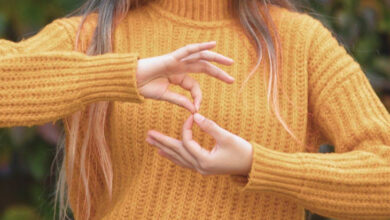 Görselde, hardal sarısı kalın örgü kazak giymiş bir kişi yer alıyor. Kişinin yüzü görünmüyor, ancak uzun, açık kahverengi ve uçları açık renk tonlarına sahip saçları dikkat çekiyor. Ellerini kullanarak işaret diliyle bir kelime veya ifade oluşturuyor. Arka planda yeşil ve kahverengi tonlarında bulanık bir doğal alan bulunuyor.