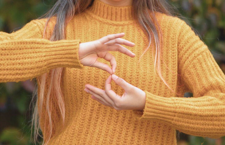 Görselde, hardal sarısı kalın örgü kazak giymiş bir kişi yer alıyor. Kişinin yüzü görünmüyor, ancak uzun, açık kahverengi ve uçları açık renk tonlarına sahip saçları dikkat çekiyor. Ellerini kullanarak işaret diliyle bir kelime veya ifade oluşturuyor. Arka planda yeşil ve kahverengi tonlarında bulanık bir doğal alan bulunuyor.