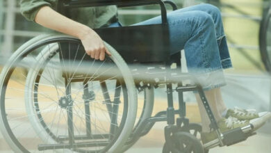 Görselde, tekerlekli sandalyede oturan bir kişi yer alıyor. Kişi, mavi kot pantolon ve yeşil spor ayakkabı giymiş. Sağ eliyle tekerlekli sandalyenin büyük tekerleğini kavrayarak hareket ettiriyor. Arka planda cam bir bariyer veya pencere yansımaları görülebilir.