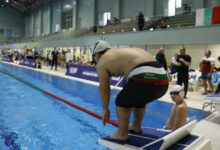Fotoğrafta, bir yüzme yarışması veya etkinliği sırasında havuz kenarında start pozisyonu alan bir sporcu görülüyor. Sporcu, yüzme başlama platformunun üzerinde eğilmiş şekilde duruyor, yüzme bonesi ve gözlük takıyor. Üzerindeki mayo Bulgaristan bayrağı renklerinde ve "Grigor Stanchev" yazısı dikkat çekiyor. Hemen yanında daha küçük bir sporcu oturuyor, gözlük ve bone takmış durumda.