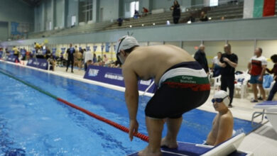 Fotoğrafta, bir yüzme yarışması veya etkinliği sırasında havuz kenarında start pozisyonu alan bir sporcu görülüyor. Sporcu, yüzme başlama platformunun üzerinde eğilmiş şekilde duruyor, yüzme bonesi ve gözlük takıyor. Üzerindeki mayo Bulgaristan bayrağı renklerinde ve "Grigor Stanchev" yazısı dikkat çekiyor. Hemen yanında daha küçük bir sporcu oturuyor, gözlük ve bone takmış durumda.