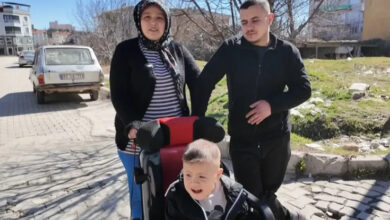 Görselde, açık havada çekilmiş bir karede üç kişi yer alıyor. Ön planda özel bir engelli tekerlekli sandalyede oturan küçük bir çocuk bulunuyor. Arkasında, çocuğun annesi olduğu düşünülen başörtülü bir kadın ve yanında genç bir adam duruyor. Arka planda apartmanlar, ağaçlar ve eski bir araç görülüyor.