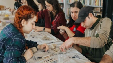 Görselde, bir grup insanın seramik veya heykel yapımıyla ilgilendiği bir atölye ortamı görülüyor. Masanın üzerinde kille şekillendirme için kullanılan aletler, su kapları ve çeşitli malzemeler bulunuyor. Ön planda kırmızı saçlı genç bir kadın ve yeşil kapüşonlu bir adam çalışıyor. Arkada ise farklı yaşlarda ve görünümlerde başka katılımcılar da sanatla uğraşıyor. Ortam samimi ve yaratıcı bir atmosfere sahip görünüyor.