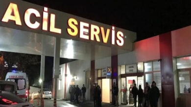 Fotoğrafta, gece vakti bir hastanenin acil servis girişi görülüyor. Büyük harflerle yazılmış "ACİL SERVİS" tabelası parlak ışıklarla aydınlatılmış. Kapının önünde birkaç kişi bekliyor, bazıları ayakta duruyor. Bir ambulans, acil servis girişinin yakınında park etmiş, mavi tepe lambası yanıyor.