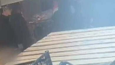 Görsel, bir market içindeki güvenlik kamerası kaydından alınmış gibi görünüyor. Ön planda raflar ve alışveriş sepetleri bulunuyor. Arka planda ise loş bir ortamda birkaç kişi yer almakta. Görüntü bulanık ve parlama nedeniyle detaylar net şekilde seçilemiyor, ancak içeride bir hareketlilik olduğu izlenimi veriyor.