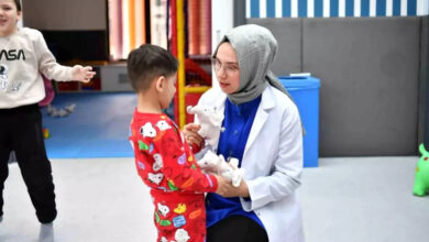 Görselde, başörtülü ve beyaz önlük giymiş bir kadın, küçük bir çocukla etkileşimde bulunuyor. Kadın, çocuğa beyaz bir oyuncak göstererek onunla iletişim kuruyor. Çocuk kırmızı pijama giymiş ve dikkatlice oyuncağa bakıyor. Arka planda bir başka çocuk oyun oynuyor. Ortam, rengarenk ve çocuk dostu bir alan gibi görünüyor; bu da bir eğitim veya terapi merkezinde olabileceklerini düşündürüyor.