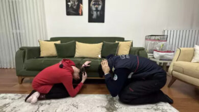 Görselde, bir ev ortamında iki kişi diz çökerek başlarını koruma pozisyonuna almış halde görülüyor. Kırmızı kıyafetli bir kadın ve itfaiye üniforması giymiş bir görevli, olası bir afet anında alınması gereken önlemleri uygulamalı olarak gösteriyor. Arkada yeşil bir koltuk, dekoratif yastıklar ve duvarda sanatsal tablolar bulunuyor. Bu görüntü, itfaiye ekiplerinin zihinsel engelli bir bireye afet eğitimi verdiği bir anı yansıtıyor.