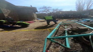 Görselde, bir olay yeri görülüyor. Sarı güvenlik şeridiyle çevrelenmiş bir alan bulunuyor. Olay yeri inceleme ekipleri veya görevliler, yerde yatan bir kişiye müdahale ediyor. Arka planda eski taş yapılar, bazı eşyalar ve bir iş makinesi dikkat çekiyor. Görüntü, kaza veya benzeri bir olayın yaşandığı bir alanı yansıtıyor.