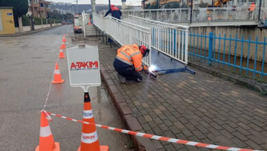 Görselde, Kocaeli Büyükşehir Belediyesi ekipleri tarafından Yahya Kaptan Mahallesi Akarca Köprüsü'nde yapılan engelli rampasının tamamlanma süreci görülüyor. İşçiler, güvenlik önlemleri alınmış bir alanda kaynak yaparak rampanın montajını gerçekleştiriyor. Çalışma alanı trafik konileri ve uyarı tabelaları ile çevrilmiş.