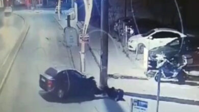 Görselde, gece vakti güvenlik kamerası tarafından kaydedilmiş bir trafik kazası anı görülüyor. Bir otomobil, kaldırıma çarpmış ve bir kişi yere savrulmuş durumda. Yakında bir elektrik direği ve park halinde araçlar bulunuyor. Görüntü, olayın şehir içi bir yolda gerçekleştiğini gösteriyor. Kaza anı ve kişinin yerde yatışı, çarpmanın şiddetli olduğunu düşündürüyor.