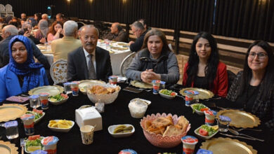 Görselde, bir iftar programında yuvarlak bir masada oturan insanlar görülüyor. Masada pide, salata, yoğurt, turşu ve çeşitli yemekler bulunuyor. Katılımcılar arasında takım elbiseli bir erkek ve başörtülü ile açık saçlı kadınlar yer alıyor. Arka planda başka misafirler de yemeklerini bekliyor. Mekân loş ışıklarla süslenmiş, sıcak bir atmosfer yaratılmış. Etkinlik, engelli bireyler ve aileleri için düzenlenen bir iftar programına ait.