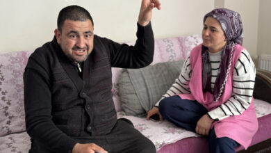 Görselde, bir kanepe üzerinde oturan iki kişi görülüyor. Soldaki adam koyu renk kıyafetler giymiş, sakallı ve elinde bir cep telefonu tutuyor. Sağ eliyle yukarıyı işaret ederken yüzünde konuşkan ve canlı bir ifade var. Sağdaki kadın ise başörtülü, pembe bir yelek giymiş ve onu dikkatlice dinlerken hafifçe gülümsüyor.