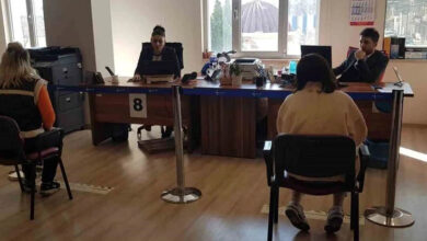 Fotoğrafta bir kamu kurumu veya ofis ortamı görülüyor. Önde iki kişi sırtı dönük şekilde sandalyelerde otururken, karşılarında masa arkasında çalışan iki görevli bulunuyor. Masada "8" numarası yer alıyor ve mavi bir şerit çekilmiş. Görevlilerden biri bilgisayar başında çalışırken diğeri bir evrakla ilgileniyor. Arka planda pencere, bir takvim ve çeşitli ofis malzemeleri bulunuyor.