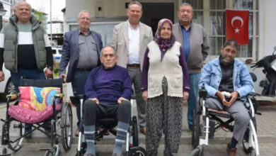 Görselde, bir grup insan bir muhtarlık binasının önünde poz veriyor. Ön sırada iki kişi tekerlekli sandalyede otururken, yanlarında başörtülü yaşlı bir kadın ayakta duruyor. Arkada ise dört erkek ayakta, gülümseyerek kameraya bakıyor. Sağ tarafta bir Türk bayrağı asılı. Yan tarafta boş bir tekerlekli sandalye bulunuyor. Fotoğraf, engelli bireylere yönelik bir destek veya dayanışma etkinliğini yansıtıyor.