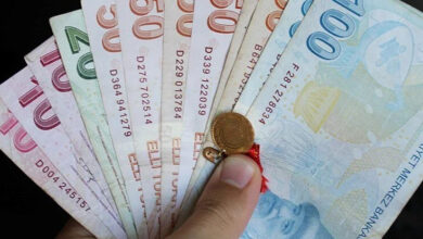 Görselde, bir elin tuttuğu farklı değerlerde Türk Lirası banknotları ve bir adet altın görünüyor. Banknotlar arasında 10, 20, 50 ve 100 TL bulunuyor. Altın, küçük bir kurdeleye iliştirilmiş şekilde parmakla tutuluyor. Arka plan koyu renkte ve detaylar net bir şekilde görülüyor.