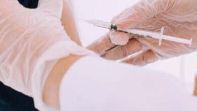 Fotoğrafta, bir sağlık çalışanı steril eldivenler giymiş halde bir kişiye aşı yapıyor. Enjeksiyon, kişinin üst kol bölgesine uygulanıyor. Sahne, steril bir ortamda gerçekleşiyor ve sağlık prosedürüne uygun bir şekilde yürütülüyor.