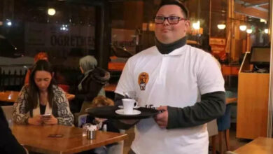 Görselde, bir kafede çalışan genç bir birey tepside kahve fincanı taşıyor. Beyaz bir tişört ve altında koyu renk kıyafetler giymiş, gözlük takıyor ve gülümseyerek hizmet veriyor. Arka planda kafe içinde oturan müşteriler, masalar ve sıcak bir ortam görülüyor. Engelsiz Kafe’de çalışan bireylerin toplumsal hayata katılımını destekleyen bir girişim olduğu anlaşılıyor.