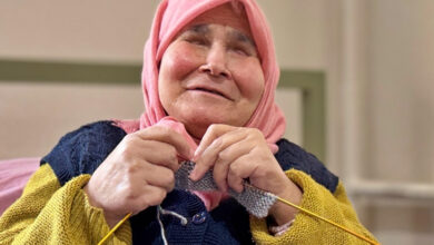 Görselde, pembe başörtüsü takan yaşlı bir kadın, elinde şiş ve iplerle örgü örüyor. Üzerinde sarı kollu kalın bir kazak ve lacivert bir yelek bulunuyor. Yüzünde hafif bir gülümseme var ve gözleri tamamen kapalı gibi görünüyor. Arka plan nötr tonlarda ve iç mekânda çekilmiş bir fotoğraf izlenimi veriyor.