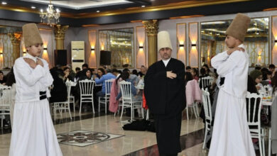 Fotoğrafta, bir salonda semazen gösterisi gerçekleştiriliyor. İki semazen beyaz kıyafetleri ve büyük kahverengi sikke şapkalarıyla gösteriye hazırlanıyor. Ortalarında siyah cübbeli bir kişi bulunuyor. Arka planda yuvarlak masalarda oturan davetliler iftar programına katılıyor. Salon, şık avizeler, aynalar ve altın rengi detaylarla süslenmiş.