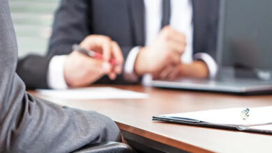 Görselde, bir iş görüşmesi ya da resmi bir toplantı ortamı olduğu anlaşılan bir sahne yer almaktadır. Ön planda tekerlekli sandalyede oturan bir bireyin kolu ve sandalyenin bir kısmı görülmektedir. Kişi, takım elbise giymiştir. Masanın üzerinde bir kalem ve evraklar bulunmaktadır. Arka planda ise takım elbiseli başka bir kişi daha vardır; bu kişi ellerini kullanarak konuşuyor gibi görünmektedir.