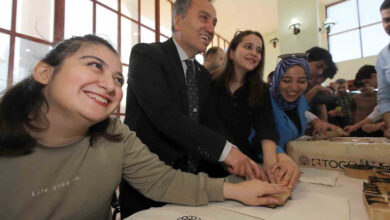 Görselde, bir grup insan geleneksel el sanatları etkinliğinde bir araya gelmiş. Ön planda genç bir kadın gülümseyerek elini bez üzerine bastırıyor. Yanında takım elbiseli bir adam ve birkaç kişi daha aynı işlemi yaparken mutlu görünüyor. Masada Tokat yazması baskıları için kullanılan ahşap kalıplar bulunuyor. Arka planda daha fazla katılımcı çalışıyor.