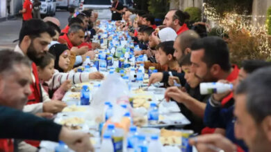 Görselde, uzun bir masada açık havada topluca yemek yiyen insanlar görülüyor. Masanın üzerinde su, ayran ve çeşitli yemeklerin bulunduğu tepsiler yer alıyor. Katılımcılar arasında çocuklar ve yetişkinler var, bazı kişiler Kızılay yeleği giymiş durumda. Arka planda ışık süslemeleri ve park edilmiş araçlar dikkat çekiyor.