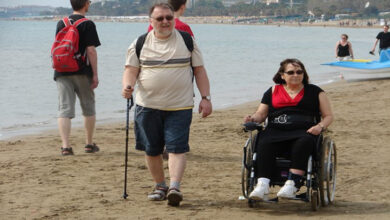 Fotoğrafta, deniz kenarında kumsalda yürüyen ve tekerlekli sandalyeyle ilerleyen iki kişi görülüyor. Adam, kısa pantolon ve tişört giymiş, elinde bir baston taşıyor. Kadın ise tekerlekli sandalyesiyle kumsalda hareket ediyor ve rahat görünüyor. Arka planda denizde yüzen ve sahilde yürüyen başka insanlar, oteller ve plajın genel atmosferi dikkat çekiyor.