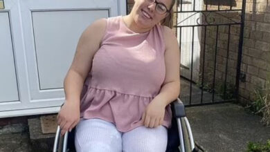 Fotoğrafta, bir kadın tekerlekli sandalyede oturuyor ve gülümsüyor. Gözlük takıyor ve rahat kıyafetler giymiş. Arkasında bir kapı ve küçük bir bahçe alanı bulunuyor.