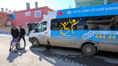 Görselde, Van Büyükşehir Belediyesi'ne ait bir engelli hizmet aracı görülüyor. Aracın üzerinde "Sosyal Hizmetler Daire Başkanlığı" ve "Engelleri Hep Beraber Aşalım" yazıları yer alıyor. Ayrıca, iletişim hattı olarak "ALO Engelli Hizmet 444 44" numarası bulunuyor. Aracın yanında, bir kişi tekerlekli sandalye ile yürürken yanında bir başka kişi ona eşlik ediyor. Arka planda renkli evler ve açık bir hava görülüyor.